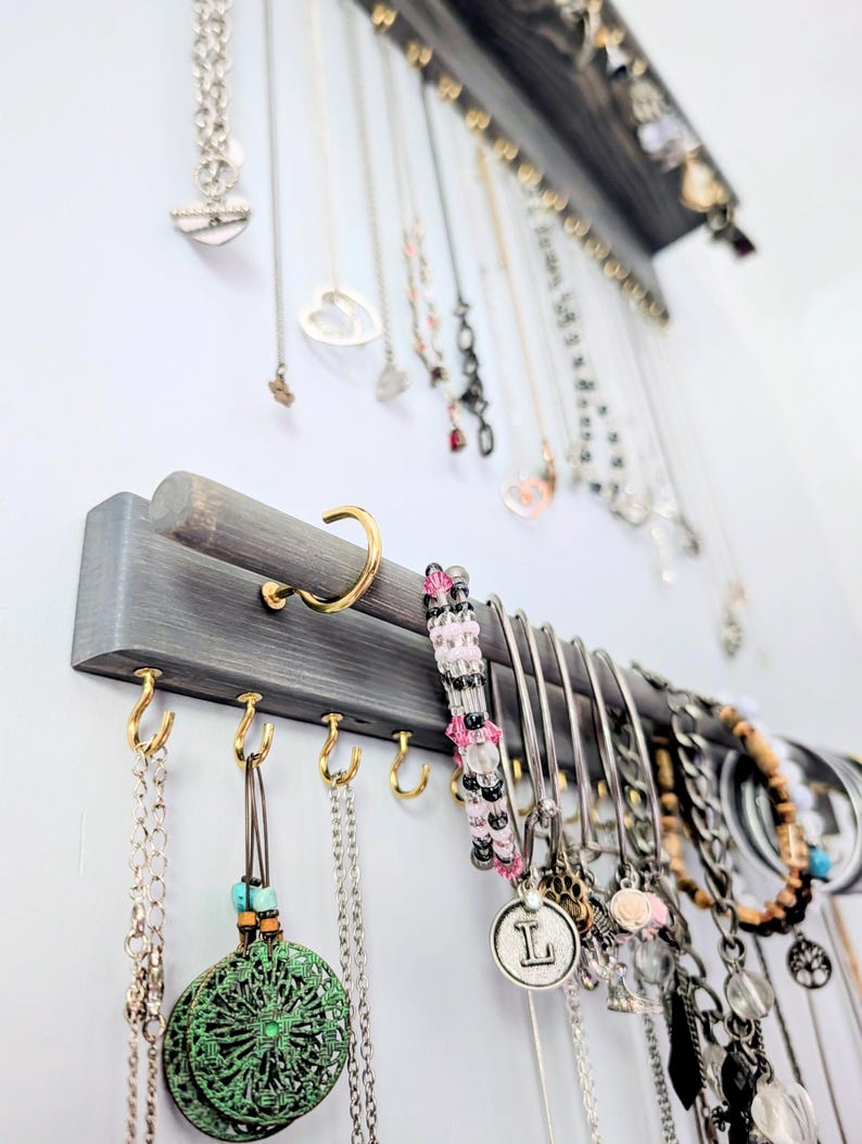 Puede incluir: Un organizador de joyas de madera gris con m&uacute;ltiples collares, pulseras y pendientes colgando. El organizador est&aacute; montado en una pared blanca y presenta ganchos dorados y una barra curva para colgar joyas.