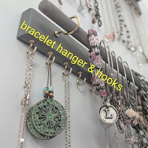 Puede incluir: Un colgador de pulseras de madera gris con ganchos dorados. El texto "bracelet hanger & hooks" est&aacute; escrito en amarillo. Varias pulseras y collares cuelgan de los ganchos.