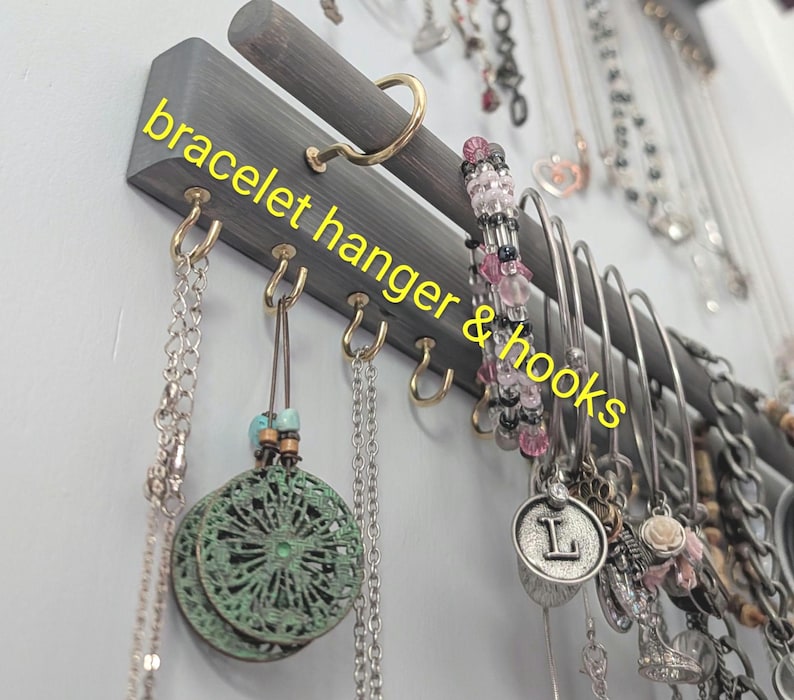 Puede incluir: Un colgador de pulseras de madera gris con ganchos dorados. El texto "bracelet hanger & hooks" est&aacute; escrito en amarillo. Varias pulseras y collares cuelgan de los ganchos.