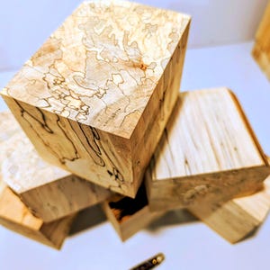 May include: A collection of five wooden blocks featuring light tan and cream tones with dark, intricate spalted grain patterns. The blocks are arranged in a stacked formation, showcasing natural wood textures and unique organic lines on each surface.
