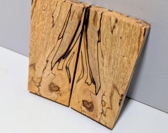 Puede incluir: Dos bloques rectangulares de madera veteada con un color base bronceado claro, con prominentes vetas orgánicas de color marrón oscuro a negro y patrones de grano intrincados. Las superficies son lisas y muestran nudos naturales.