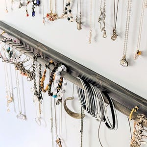 Puede incluir: Un organizador de joyas de madera gris con una barra para colgar pulseras y una barra para collares. Varios collares y pulseras en plata, oro y otros colores se exhiben en el organizador contra una pared blanca.