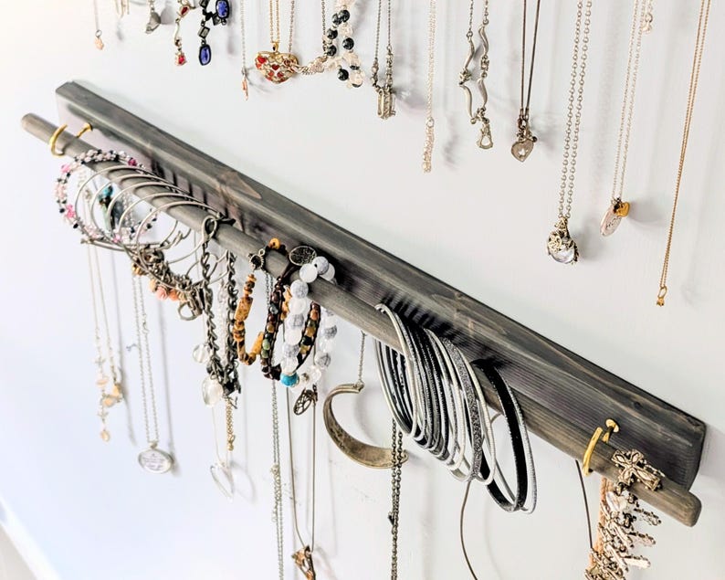 Puede incluir: Un organizador de joyas de madera gris con una barra para colgar pulseras y una barra para collares. Varios collares y pulseras en plata, oro y otros colores se exhiben en el organizador contra una pared blanca.