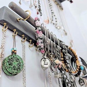 Puede incluir: Un organizador de joyas de madera gris con ganchos dorados que exhibe collares, pulseras y pendientes. Las joyas incluyen cadenas de plata, pulseras de cuentas y colgantes con la letra "L". El organizador est&aacute; montado en una pared blanca.