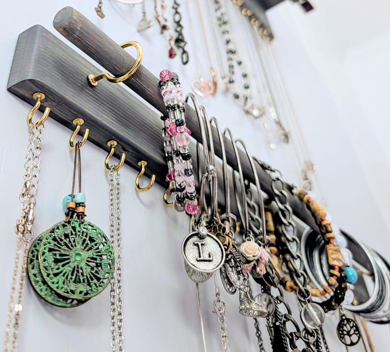 Puede incluir: Un organizador de joyas de madera gris con ganchos dorados que exhibe collares, pulseras y pendientes. Las joyas incluyen cadenas de plata, pulseras de cuentas y colgantes con la letra "L". El organizador est&aacute; montado en una pared blanca.