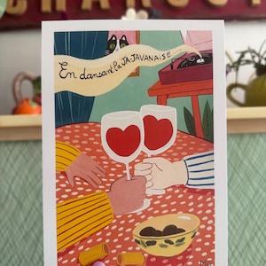 Puede incluir: Una lámina de arte rectangular con una ilustración de dos personas brindando con copas de vino que contienen corazones rojos. La mesa tiene un mantel de lunares rojos y blancos, un cuenco de aceitunas y dos corchos. El texto dice: En dansant la JA-JAVANAISE.