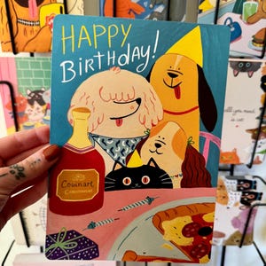 Può includere: Un biglietto di auguri con la scritta "Happy Birthday!" presenta illustrazioni di cani, un gatto nero, una bottiglia di champagne e una pizza. Il biglietto ha uno sfondo blu ed è esposto davanti ad altri biglietti.