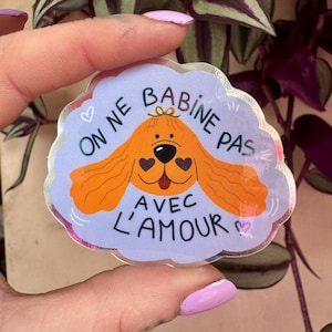 Peut inclure: Un autocollant transparent avec un dessin de chien de dessin animé. Le chien est orange avec des yeux en forme de cœur noirs et un nez noir. Le texte français "ON NE BABINE PAS AVEC L'AMOUR" est écrit en blanc.