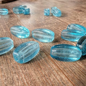 May include: Several translucent, light blue glass beads in different shapes, including oval and rectangular. The beads have a ribbed texture and are arranged on a wooden surface. These beads are suitable for jewellery making or crafting projects.