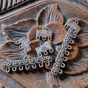 May include: Silver-coloured metal jewellery findings, including rectangular and floral-patterned connectors. The findings are displayed on a carved wooden surface with a floral design, highlighting their intricate details and potential for jewellery making.