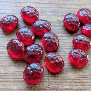 May include: A collection of translucent, red glass beads, each shaped like a strawberry. The beads have a textured surface and are scattered on a wooden surface. The beads are a deep, rich red colour.