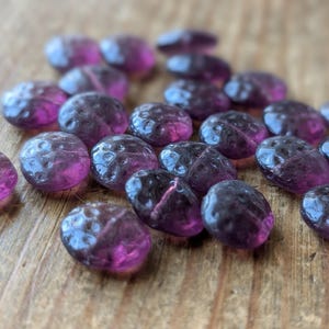 Puede incluir: Un conjunto de cuentas de vidrio translúcido morado con una superficie texturizada. Las cuentas ovaladas están dispersas sobre una superficie de madera, con un tono rosa vibrante visible dentro de cada cuenta. Las cuentas miden aproximadamente 2 cm de diámetro.