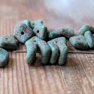May include: Close-up of turquoise textured beads with brown accents. The beads are square and cylindrical, with holes for stringing. The beads are on a wooden surface.