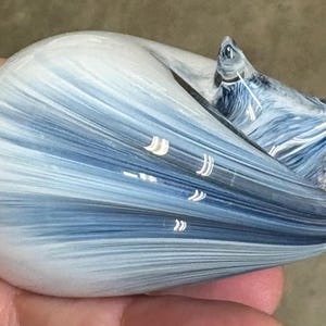 May include: A smooth, rounded glass figurine shaped like a curled animal, featuring a swirling pattern of white and deep blue streaks throughout the translucent material. The object has a polished, glossy finish and a tapered, pointed tail section.