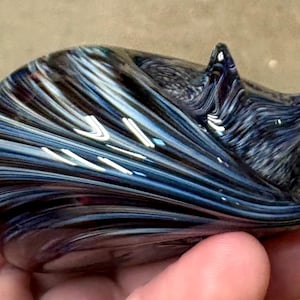 May include: A small, hand-held glass figurine shaped like a curled-up animal. The glass features a swirling pattern of deep navy blue, black, and iridescent white streaks that create a smooth, glossy finish across the entire curved surface.