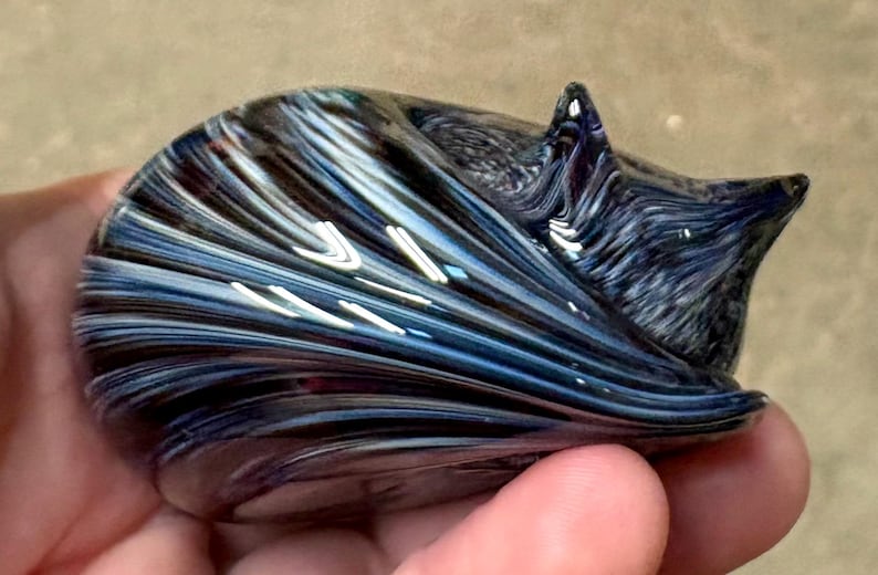 May include: A small, hand-held glass figurine shaped like a curled-up animal. The glass features a swirling pattern of deep navy blue, black, and iridescent white streaks that create a smooth, glossy finish across the entire curved surface.