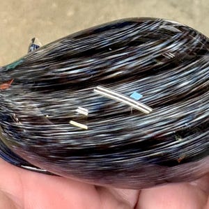 May include: A smooth, rounded glass paperweight featuring a swirling pattern of black, white, and dark blue colors. The surface has a glossy finish with subtle, embedded metallic-looking flecks and a small, pointed protrusion on one side.