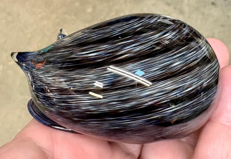 May include: A smooth, rounded glass paperweight featuring a swirling pattern of black, white, and dark blue colors. The surface has a glossy finish with subtle, embedded metallic-looking flecks and a small, pointed protrusion on one side.