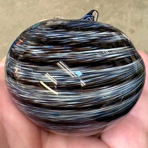 May include: A round, hand-blown glass paperweight featuring a swirling pattern of black, white, and dark blue horizontal streaks. The surface is smooth and glossy, reflecting light with a polished finish, creating a layered, marbled appearance.