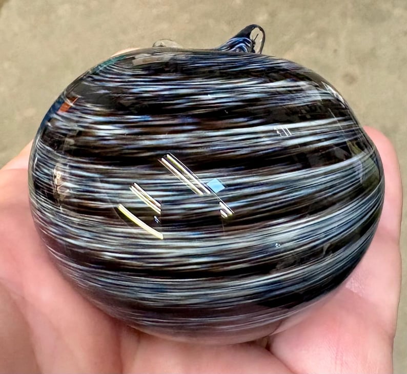 May include: A round, hand-blown glass paperweight featuring a swirling pattern of black, white, and dark blue horizontal streaks. The surface is smooth and glossy, reflecting light with a polished finish, creating a layered, marbled appearance.