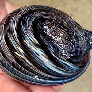 May include: A smooth, rounded glass paperweight featuring a swirling pattern of deep black, dark blue, and bright white streaks. The glass has a polished, reflective surface with a small, pointed protrusion on one side, creating a dynamic, fluid appearance.