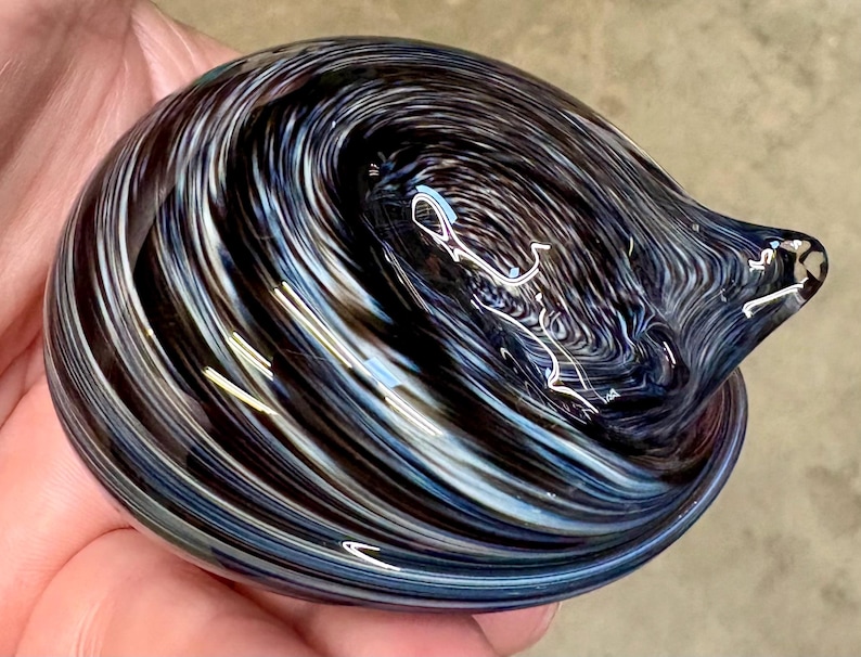 May include: A smooth, rounded glass paperweight featuring a swirling pattern of deep black, dark blue, and bright white streaks. The glass has a polished, reflective surface with a small, pointed protrusion on one side, creating a dynamic, fluid appearance.