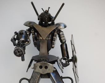 May include: A metal robot sculpture made from recycled materials. The robot is holding a gun in one hand and a shield in the other. The robot is standing next to a can of Coca-Cola.