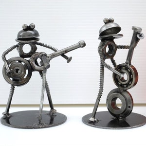 May include: Two metal frog sculptures playing musical instruments. One frog plays a guitar, the other a trumpet. The sculptures are made from nuts, bolts, and other metal hardware. A red soda can is next to the sculptures.