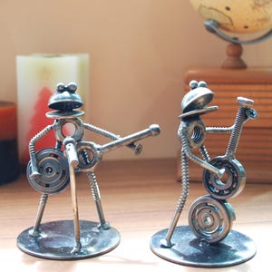 May include: Two metal frog sculptures playing musical instruments. One frog holds a microphone and a guitar, while the other plays a guitar. The sculptures are made of nuts, bolts, and washers, and are mounted on circular bases.