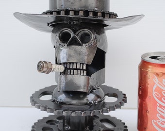May include: A metal skull sculpture wearing a hat and smoking a spark plug cigar. The sculpture is made of metal gears and other metal parts. A Coca-Cola can is next to the sculpture.
