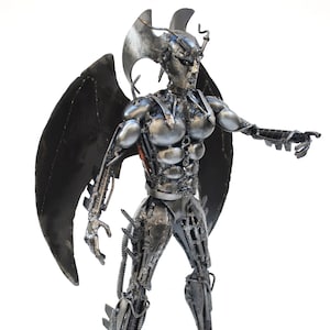 May include: A silver and black metal sculpture of an angel with wings. The angel is made of recycled metal parts, such as screws, bolts, and nuts. The angel has a muscular body and is standing with one leg forward and one leg back. The angel is pointing with its right hand.