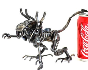 May include: A metal sculpture of an alien creature made from nuts, bolts, and other metal pieces. The sculpture is about the size of a can of soda.