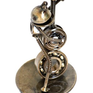 May include: A handcrafted metal sculpture of a whimsical figure, assembled from various hardware components. The sculpture features a head-like structure, two bearing wheels for a body, and a base. The metal has a tarnished, aged appearance.