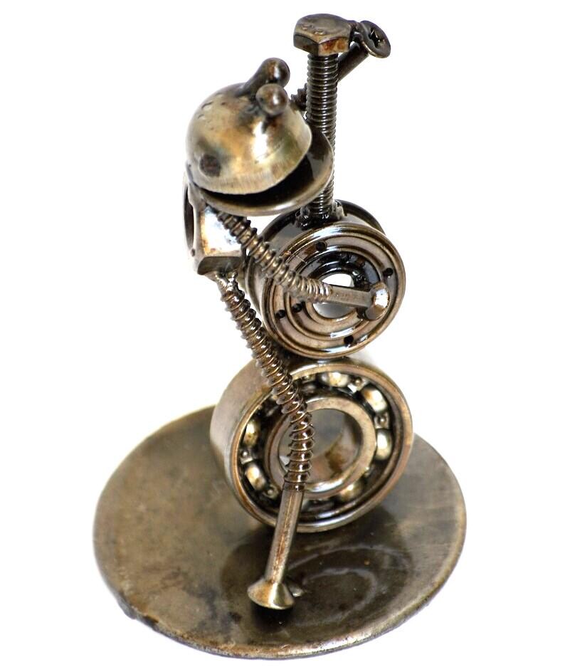 May include: A handcrafted metal sculpture of a whimsical figure, assembled from various hardware components. The sculpture features a head-like structure, two bearing wheels for a body, and a base. The metal has a tarnished, aged appearance.
