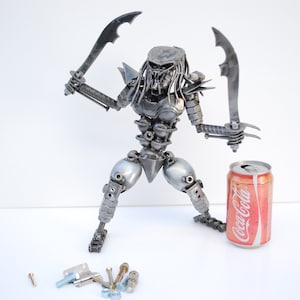 May include: A silver metal sculpture of a Predator character from the movie franchise. The sculpture is holding two swords and is standing next to a can of Coca-Cola.