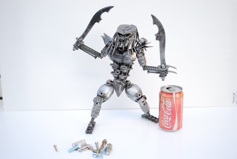May include: A silver metal sculpture of a Predator character from the movie franchise. The sculpture is holding two swords and is standing next to a can of Coca-Cola.