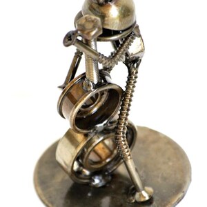 May include: A handcrafted metal sculpture of a musician playing a drum set. The sculpture is made from various metal parts, including bolts, screws, and bearings. The sculpture is on a round metal base and has a bronze finish.