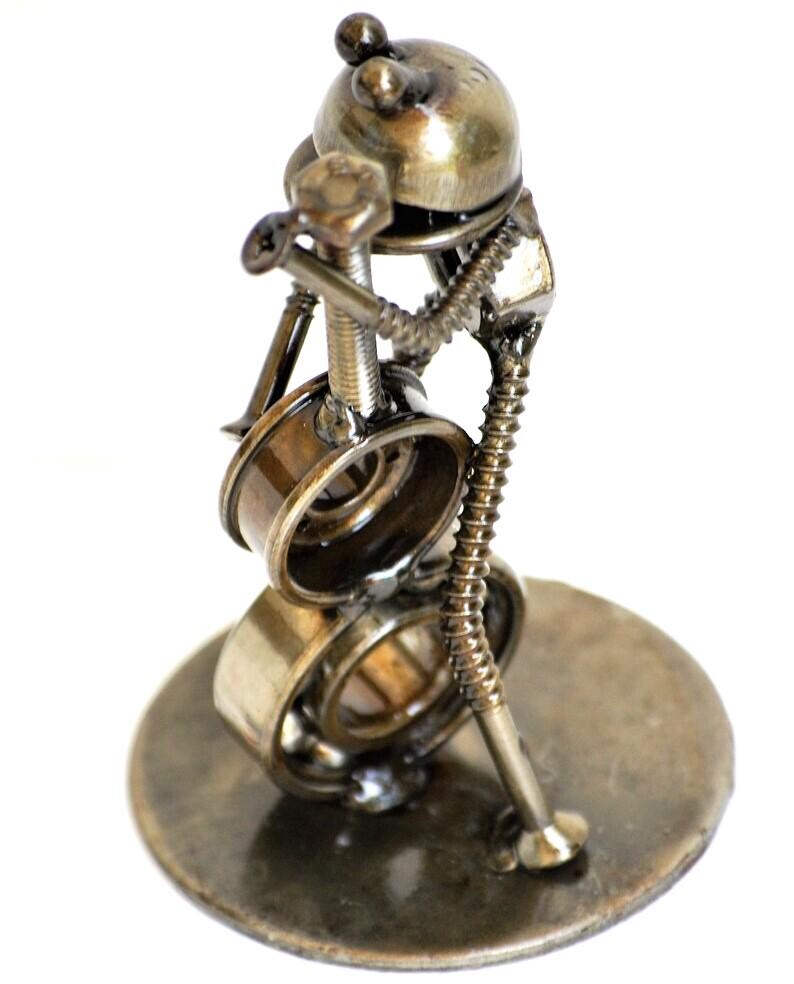 May include: A handcrafted metal sculpture of a musician playing a drum set. The sculpture is made from various metal parts, including bolts, screws, and bearings. The sculpture is on a round metal base and has a bronze finish.