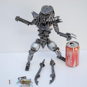 May include: A metal sculpture of a Predator from the movie franchise. The sculpture is made from recycled metal parts and is approximately 12 inches tall. The Predator is holding a weapon in its right hand and has a can of Coca-Cola next to it.