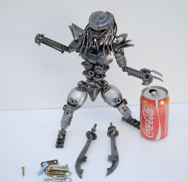 May include: A metal sculpture of a Predator from the movie franchise. The sculpture is made from recycled metal parts and is approximately 12 inches tall. The Predator is holding a weapon in its right hand and has a can of Coca-Cola next to it.