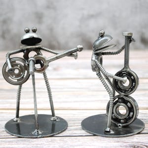 May include: Two metal frog sculptures playing musical instruments. One frog plays a banjo, the other a guitar. The sculptures are made from metal hardware and are mounted on round bases. The frogs have large eyes and are painted in a metallic silver color.
