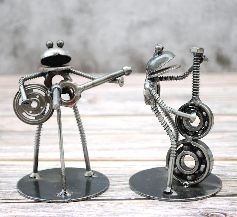 May include: Two metal frog sculptures playing musical instruments. One frog plays a banjo, the other a guitar. The sculptures are made from metal hardware and are mounted on round bases. The frogs have large eyes and are painted in a metallic silver color.
