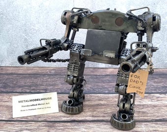 May include: A metal robot sculpture with two large guns, made from various metal components. The robot has a tag that reads "For Dad's Cave". The sculpture is displayed on a wooden surface.