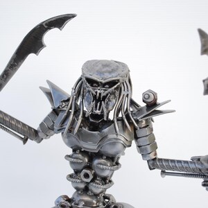 May include: A metal sculpture of a Predator from the movie franchise. The sculpture is made of recycled metal parts, including nuts, bolts, and screws. The Predator is holding two swords in its hands and has a menacing expression on its face.