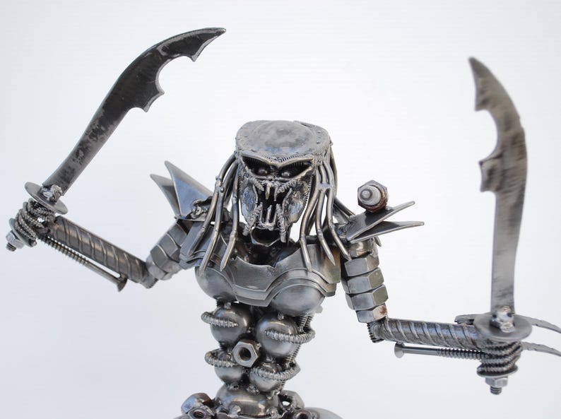 May include: A metal sculpture of a Predator from the movie franchise. The sculpture is made of recycled metal parts, including nuts, bolts, and screws. The Predator is holding two swords in its hands and has a menacing expression on its face.