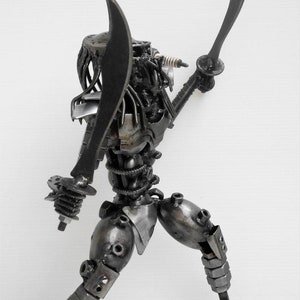 May include: A black metal sculpture of a Predator character from the movie franchise. The sculpture is made of recycled metal parts and is holding two swords.