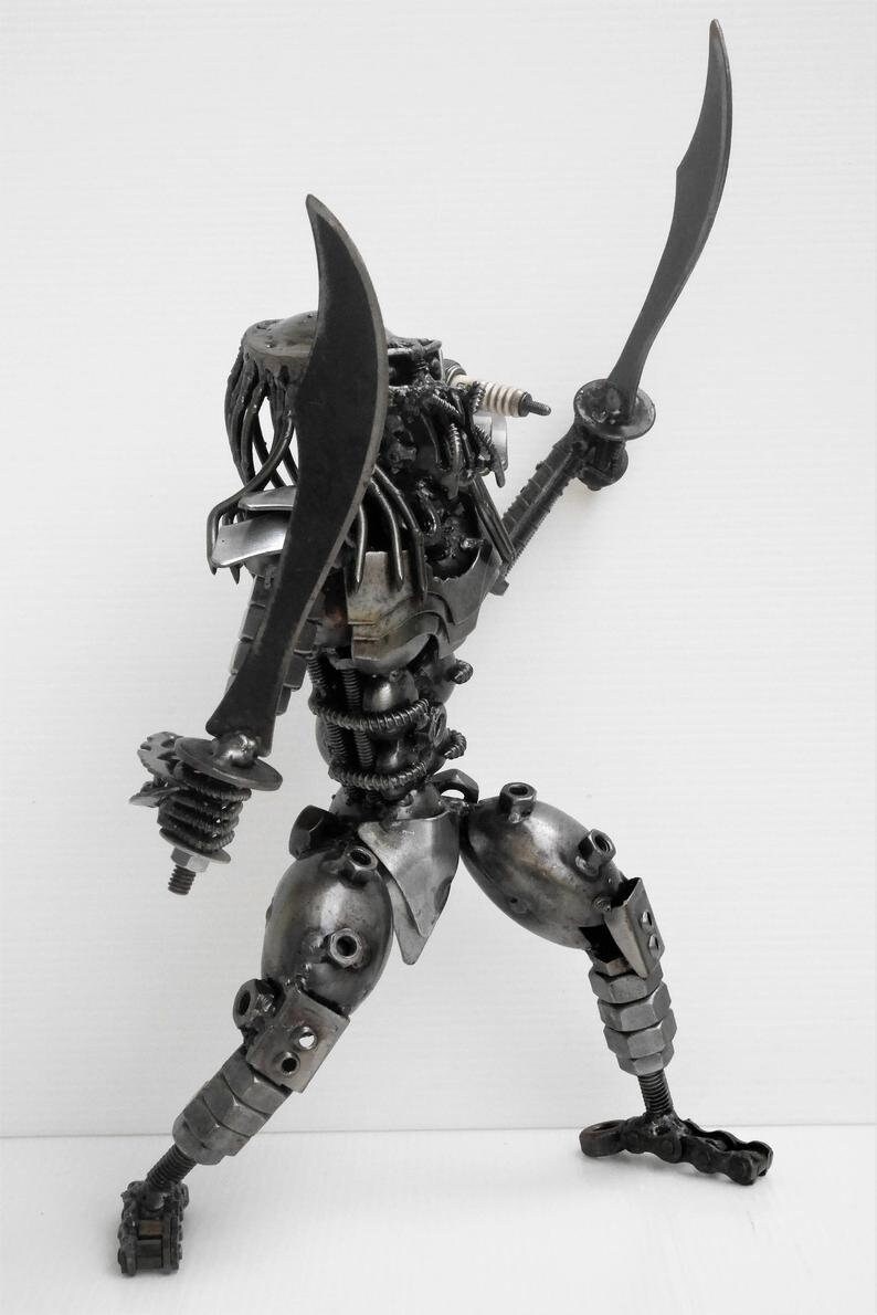 May include: A black metal sculpture of a Predator character from the movie franchise. The sculpture is made of recycled metal parts and is holding two swords.
