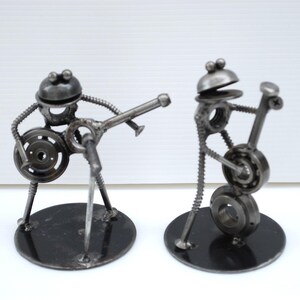 May include: Two metal sculptures of musicians. One figure plays a guitar, the other a drum. The sculptures are made from nuts, bolts, and other metal hardware. Each figure stands on a black circular base.