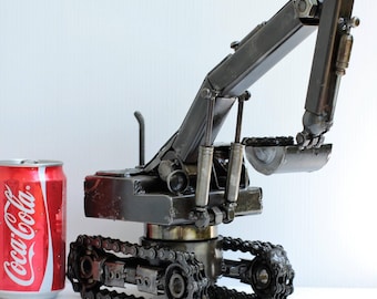 May include: A small, metal model of an excavator with a black finish. The excavator has a long arm with a bucket at the end. The excavator is sitting on a white surface next to a can of Coca-Cola.