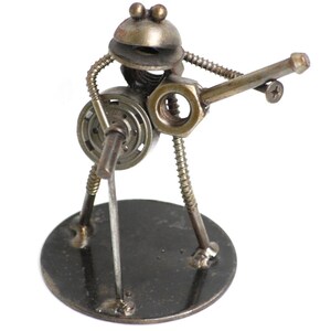 May include: A whimsical sculpture of a musician crafted from metal hardware. The figure, with a round head and body, appears to be playing a guitar. The sculpture is mounted on a circular base, showcasing a unique blend of industrial and artistic design.